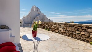 A stone-paved outdoor area with a white table and red cushions, a stone wall, and a white building with a dome in the background. - Sunset Paraportiani Rooms (Mykonos)