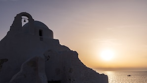 A white building with a bell tower silhouetted against a sunset, overlooking the sea. - Sunset Paraportiani Rooms (Mykonos)