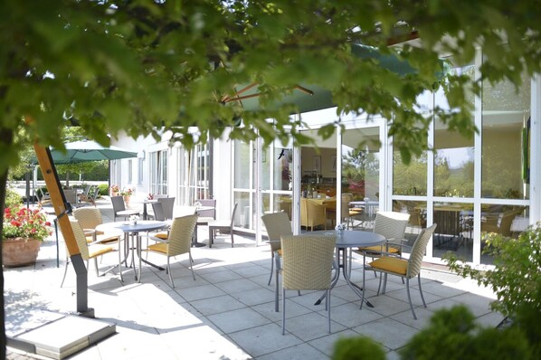 An outdoor patio with tables and chairs, large windows, and potted plants.