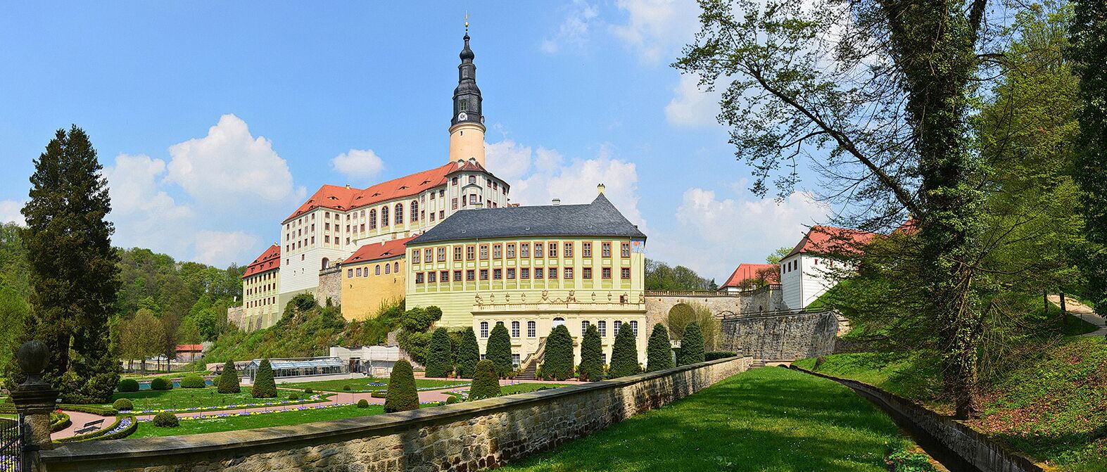 A historic building with a prominent tower, surrounded by well-maintained gardens and a stone wall.