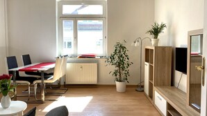 A modern living room with a dining area, a radiator, a potted plant, a floor lamp, and a TV mounted on a wooden cabinet.