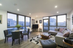 A modern living room with a sofa, chairs, a coffee table, a TV, and large windows offering a city view.