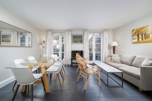 Cozy living room with a glass dining table, beige sofa, and wooden bench, featuring a fireplace and large windows.