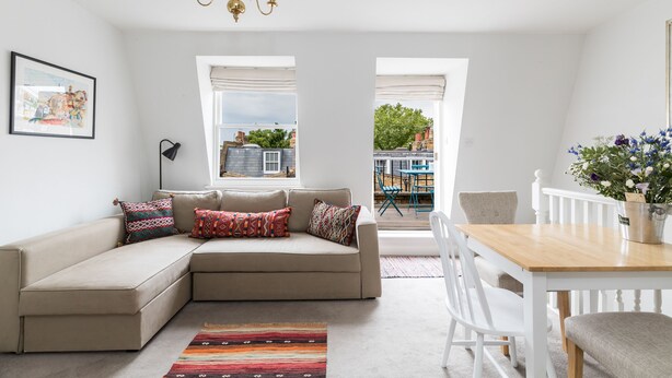 A modern living room with a beige sectional sofa, a dining table with chairs, a chandelier, and a view of the outside. - JOIVY Pimlico Flat with Terrace for 3 (London)