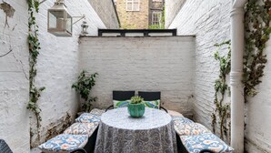 A small outdoor seating area with a round table and chairs, surrounded by white brick walls and covered with a patterned tablecloth.