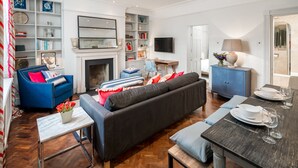 A living room with a fireplace, a sofa, a dining table set for two, and a television mounted on the wall.