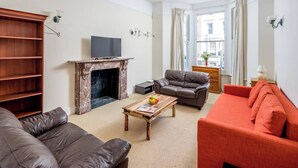 A living room with a fireplace, a television, a sofa, a red couch, and a wooden bookshelf. - JOIVY Charming 1 Bed Flat, Sleeps 4 (London)