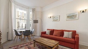 A living room with a red sofa, wooden coffee table, and patterned rug. There is a dining table with chairs and a lamp. - JOIVY Charming 1 Bed Flat, Sleeps 4 (London)