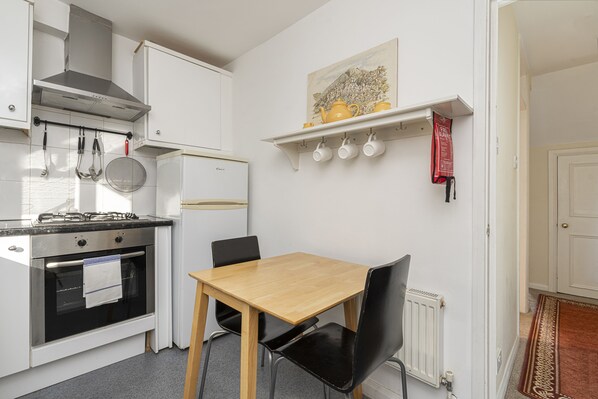 Une cuisine compacte équipée d’une cuisinière, d’un four et d’ustensiles suspendus. On y trouve une table à manger avec des chaises, et un tableau est accroché au-dessus.