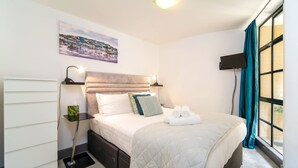 A neatly made bed with a quilt, pillows, and rolled towels, a bedside table with a lamp, a wall-mounted TV, and a window with curtains. - JOIVY Splendid 1 Bedroom Flat near St. Paul's Cathedral (London)