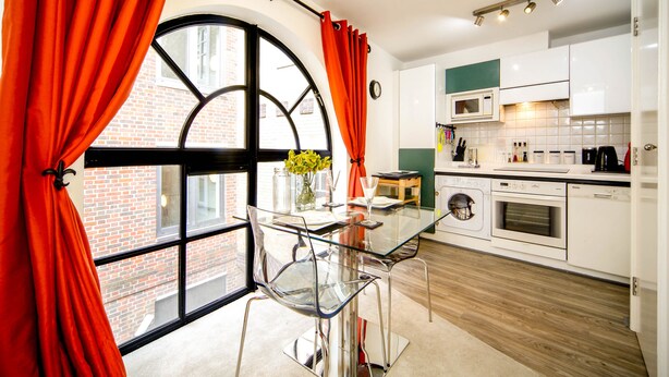 A modern kitchen with a glass dining table, red curtains, and a view of a brick building. - JOIVY Splendid 1 Bedroom Flat near St. Paul's Cathedral (London)