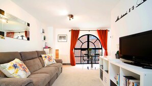 A living room with a sofa, TV, bookshelf, and a view through a window with orange curtains. - JOIVY Splendid 1 Bedroom Flat near St. Paul's Cathedral (London)