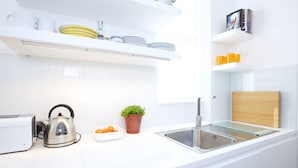 Une cuisine moderne avec des plans de travail blancs, une bouilloire en acier inoxydable, une petite plante en pot et de la vaisselle rangée avec soin sur des étagères.