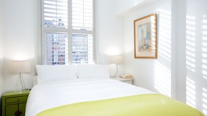 Une chambre d’hôtel avec un grand lit, une fenêtre avec des stores, une table de chevet avec une lampe et un tableau encadré au mur.