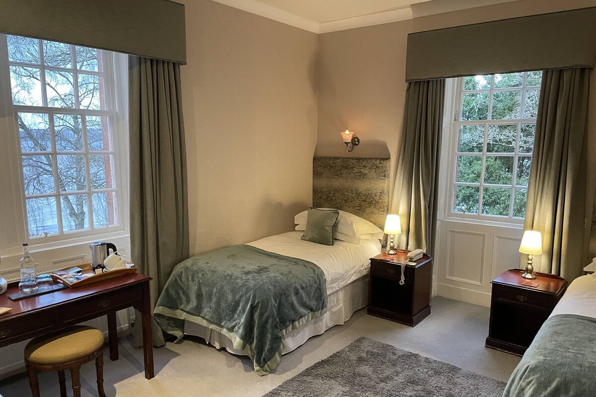 A bedroom with a bed, bedside tables, a desk, and a chair. There are windows with curtains, a rug, and a view of trees outside.