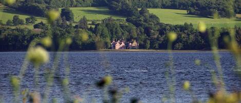 Vista de un lago con un edificio parecido a un castillo, rodeado de abundante vegetación y campos.