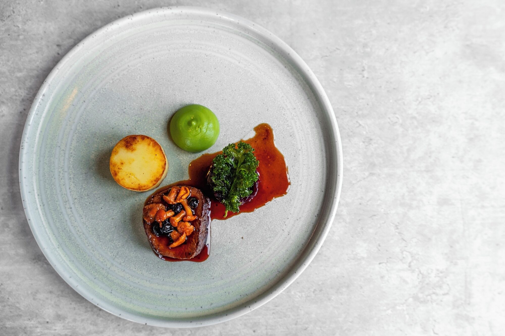 A plate with a scallop, a green fruit, and a piece of meat with mushrooms.