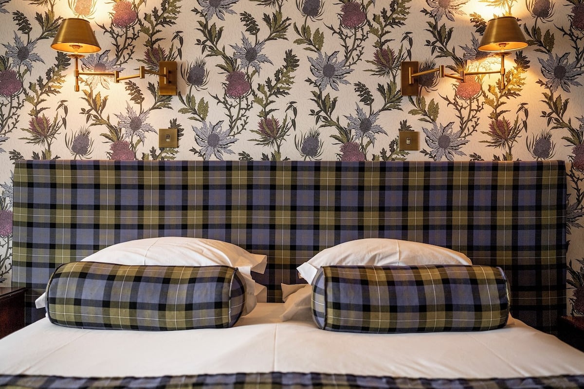 A bed with plaid bedding and pillows, a headboard with a plaid pattern, and wall-mounted lamps with floral designs.