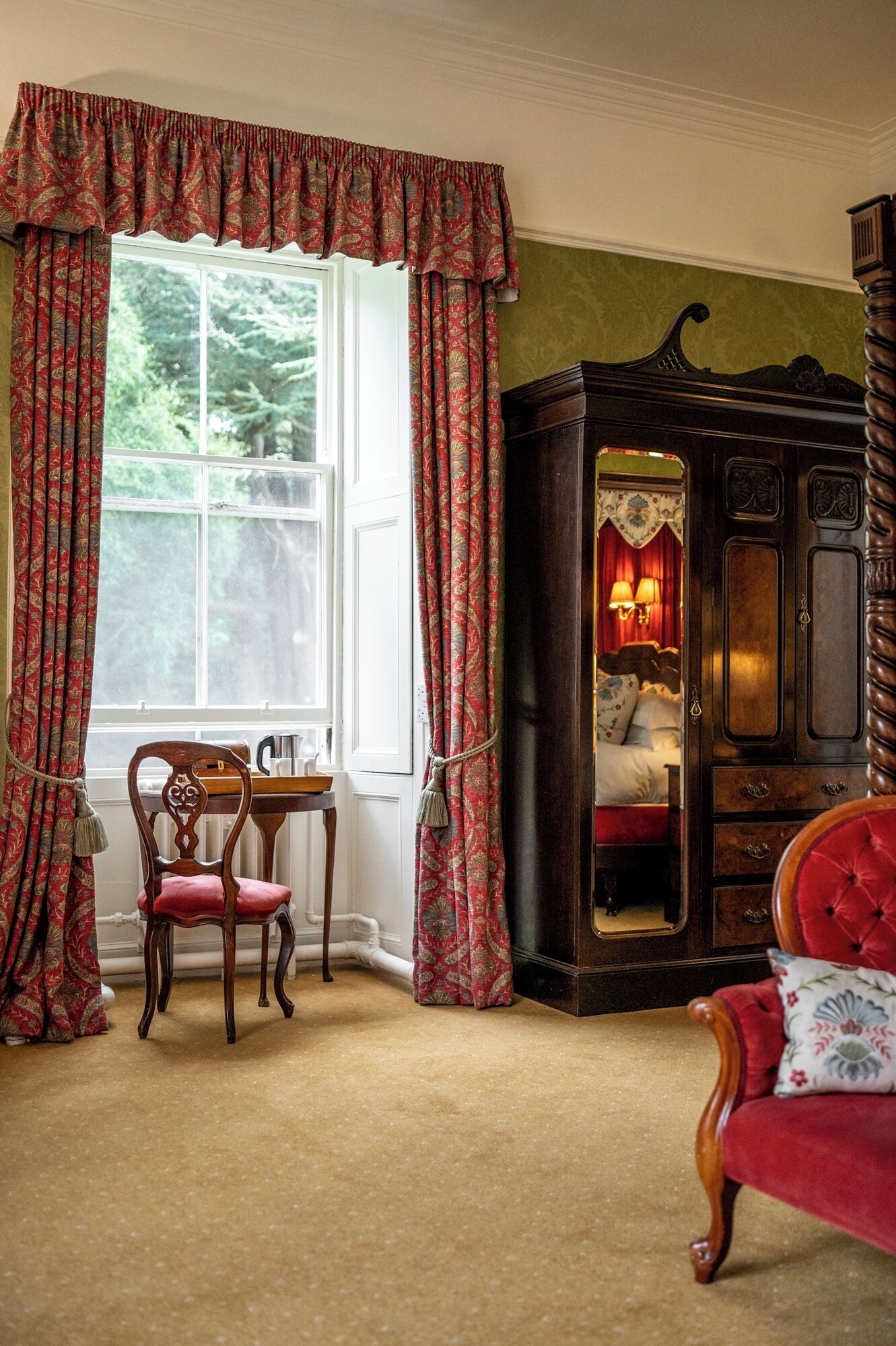 Una habitación con un ventanal grande, un armario de madera con espejo, una silla roja y una mesa con hervidor.