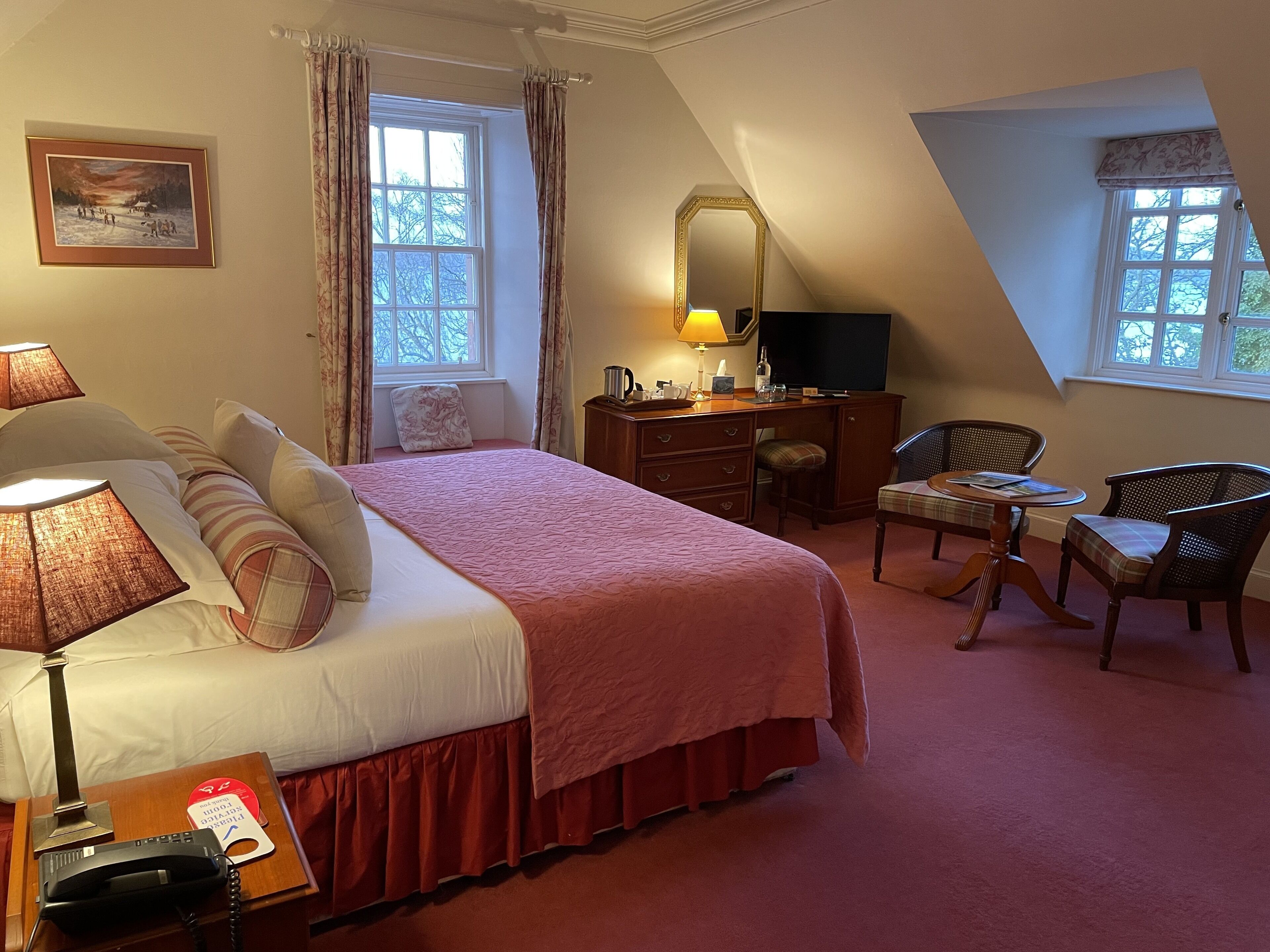 A hotel room with a large bed, a desk with a mirror, a chair, a small table, and a TV.