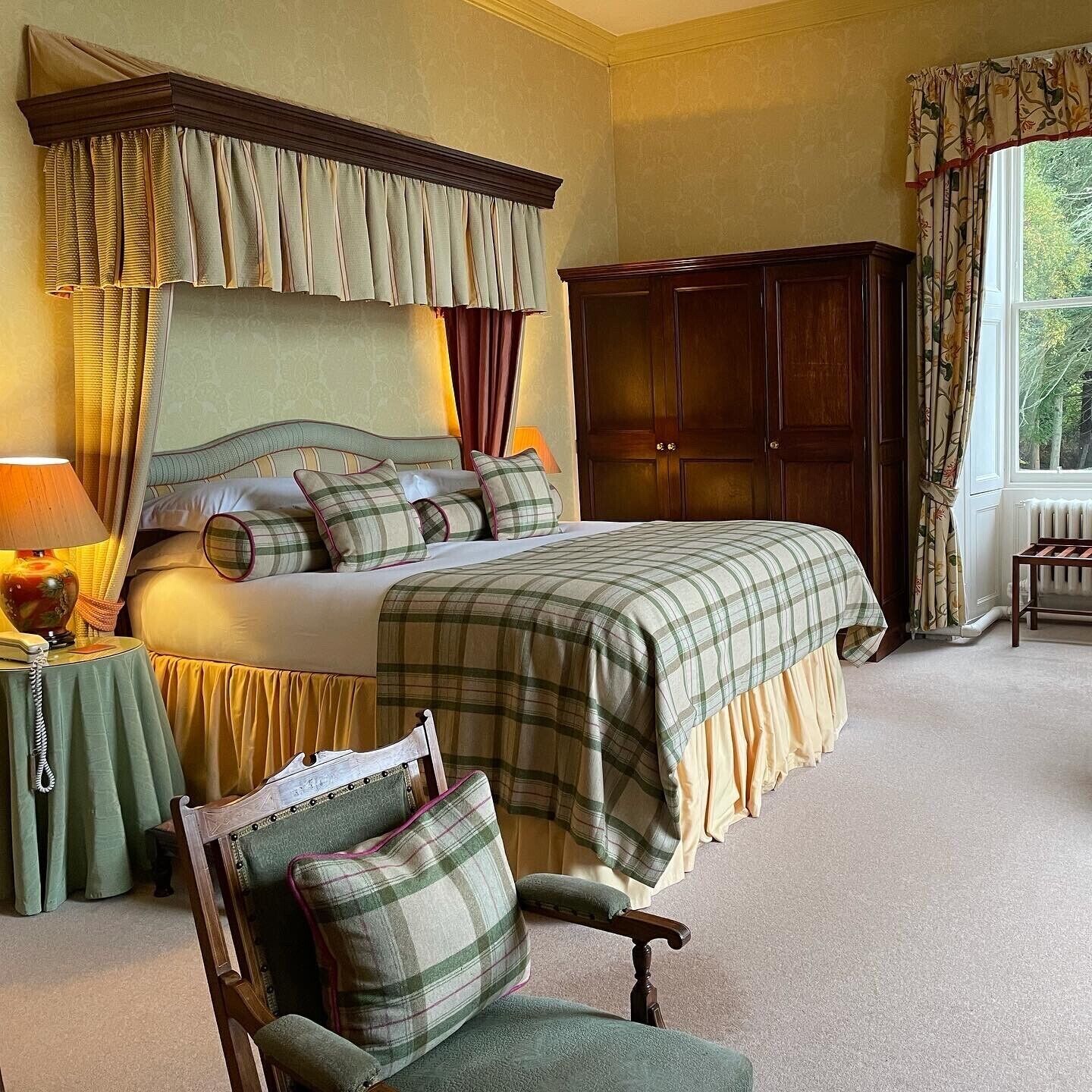 A bedroom with a large bed, a wooden wardrobe, a chair, and a table with a lamp.