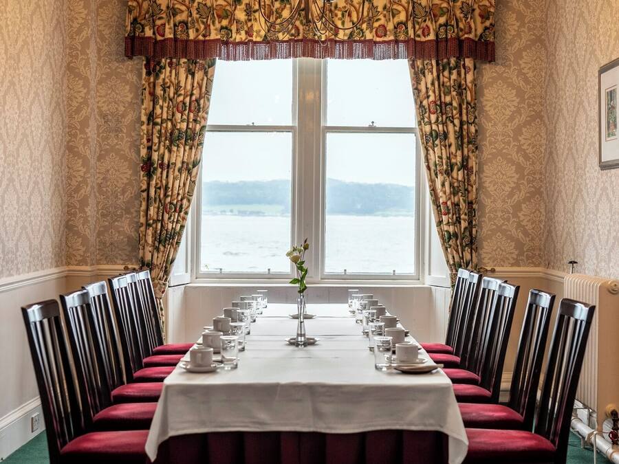 A formal dining room with a long table set for a meal, flanked by tall chairs, a chandelier, and a window with patterned curtains.
