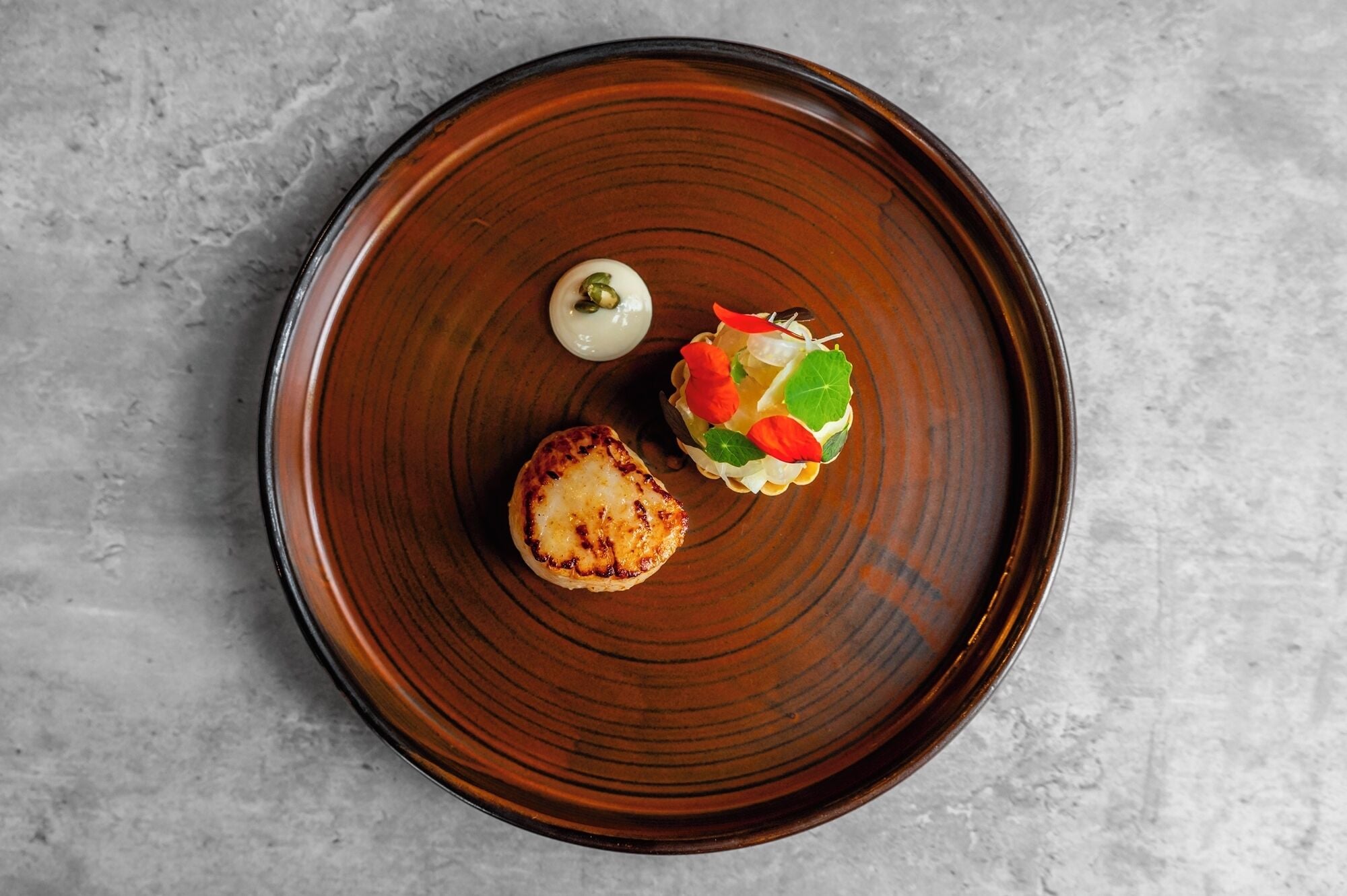 A round, dark brown plate with a grilled item and a small salad on a textured gray surface.