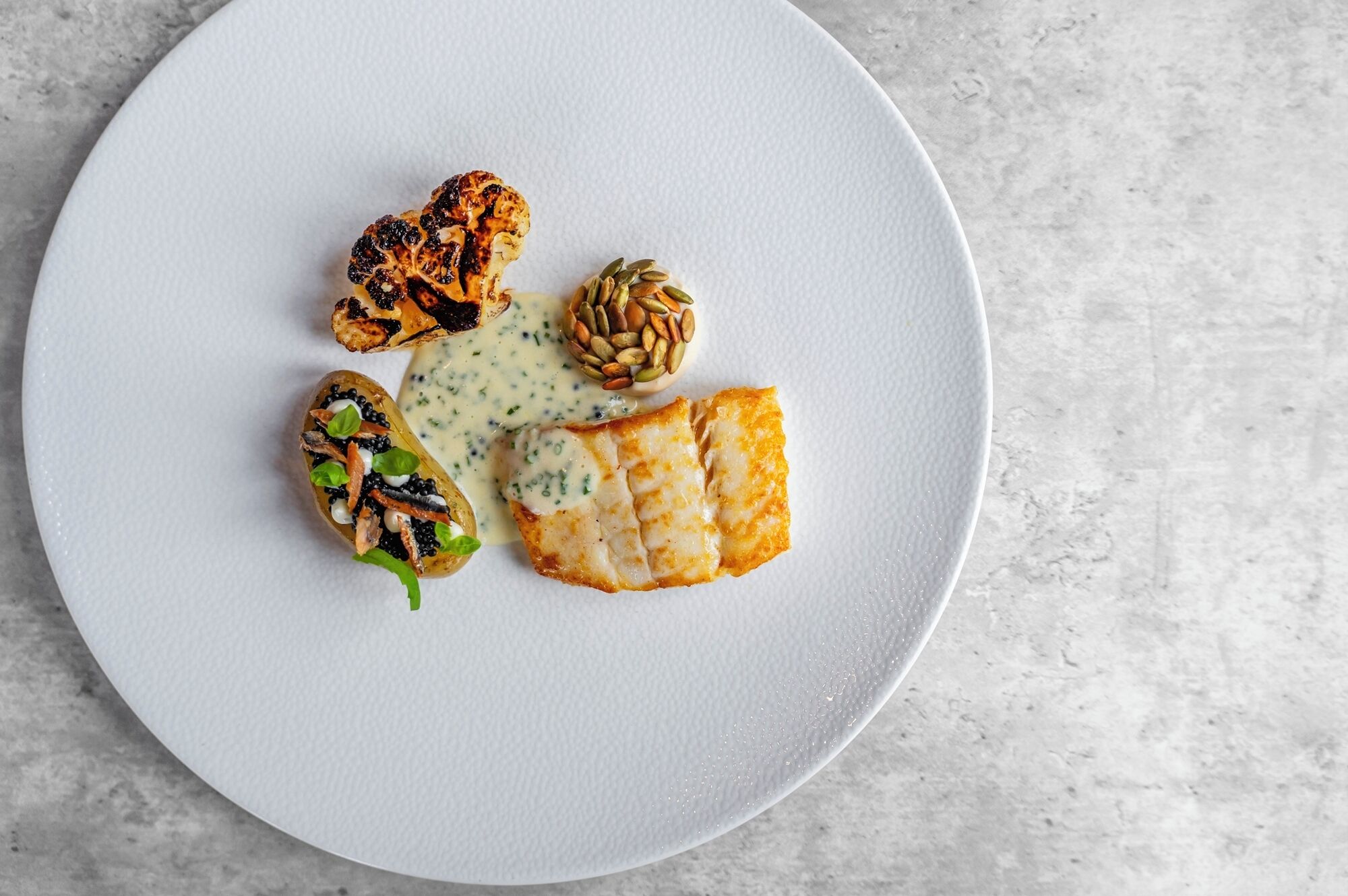 A white plate with a piece of grilled fish, a roasted cauliflower, a stuffed potato, and a small bowl of sauce.