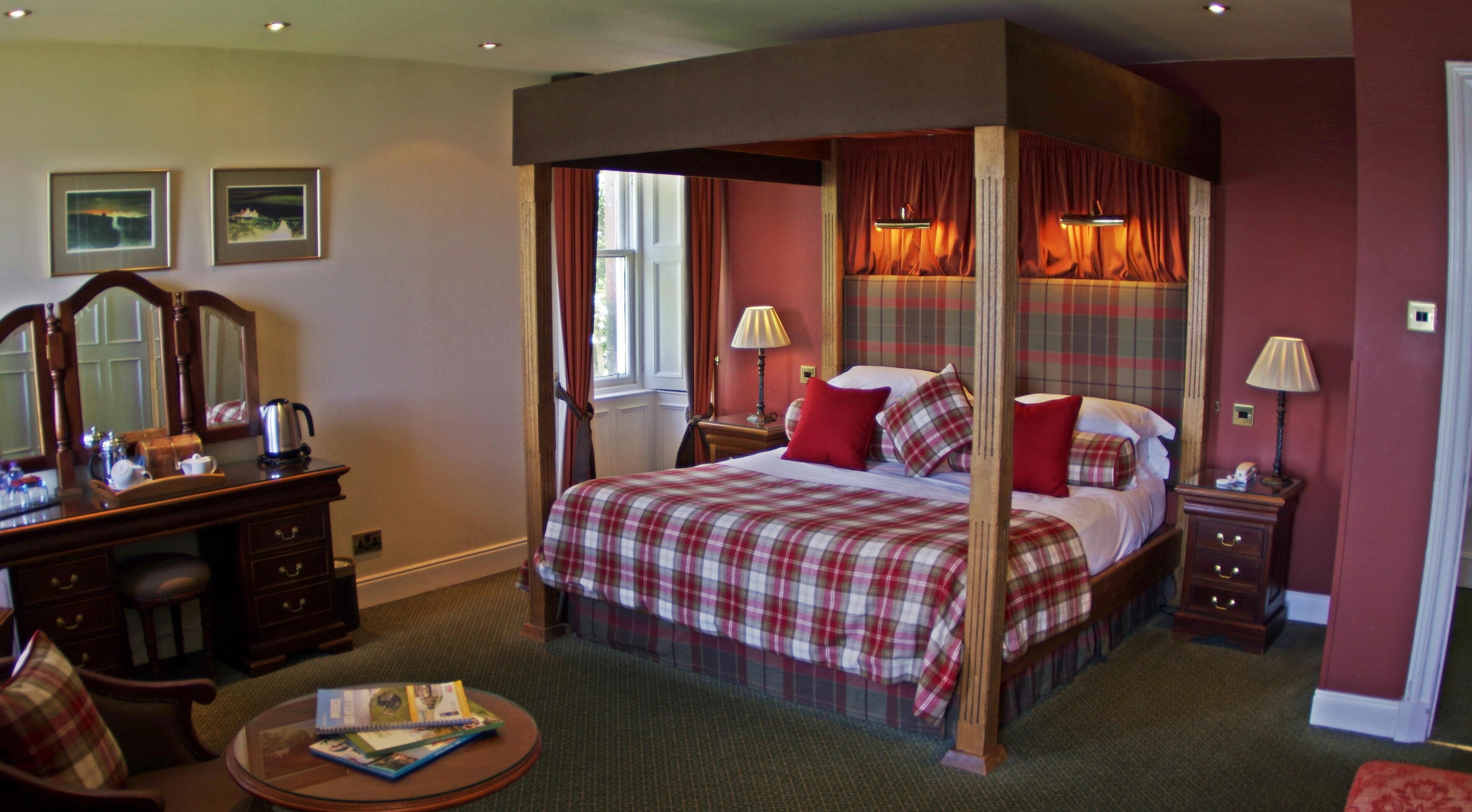 A four-poster bed with a plaid bedspread, two bedside lamps, a wooden dresser, a mirror, and a small round table with books.