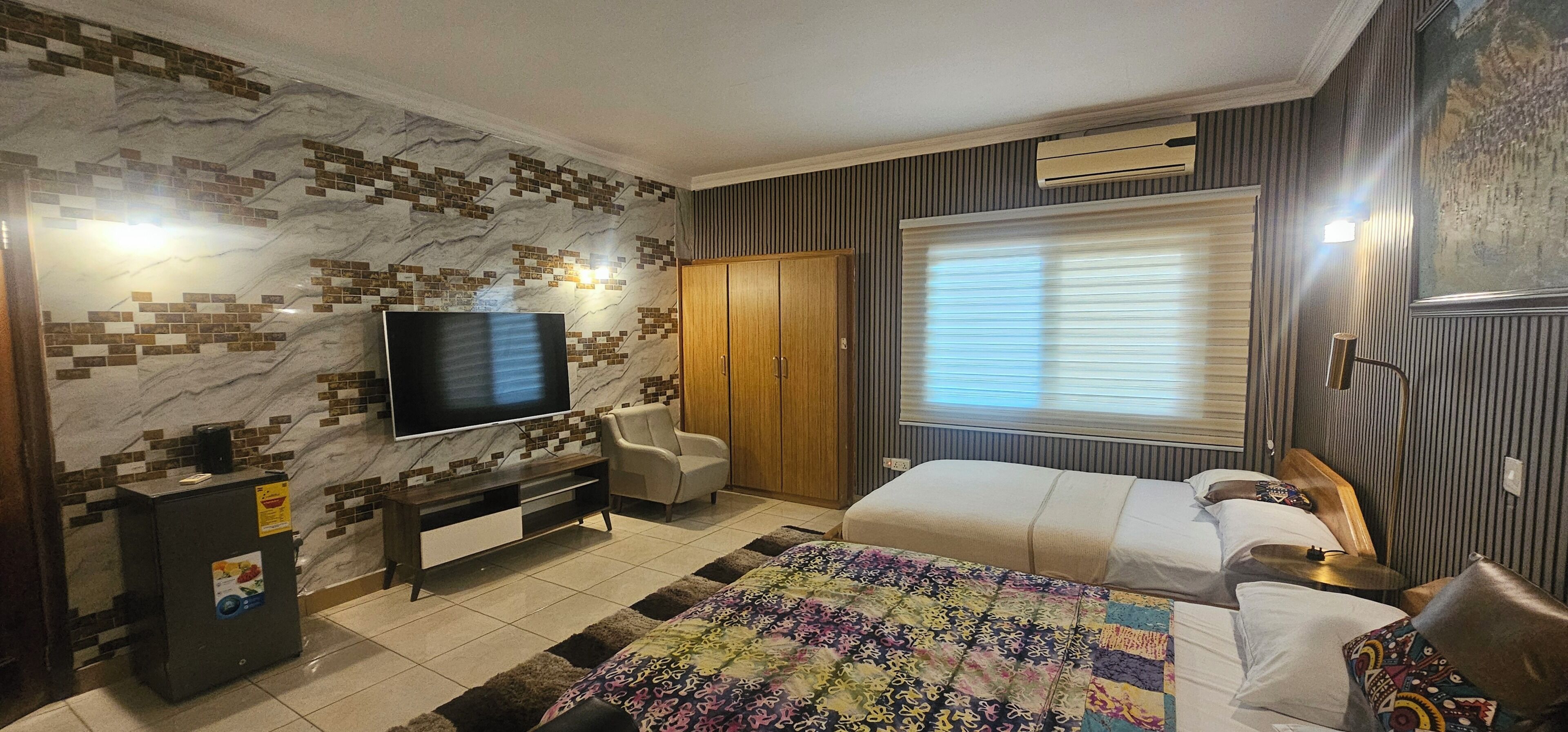 A hotel room with a TV, a bed, a chair, a wardrobe, and a window with blinds.
