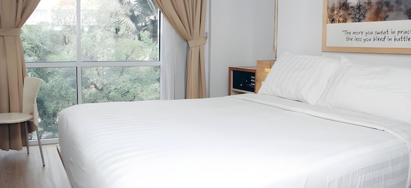 Ein ordentlich bezogenes Bett in einem Zimmer mit großem Fenster, einem Sessel und einem Nachttisch.