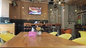 A restaurant with wooden tables, yellow chairs, a TV on the wall, and a menu board. - An Hotel Jakarta (Jakarta)
