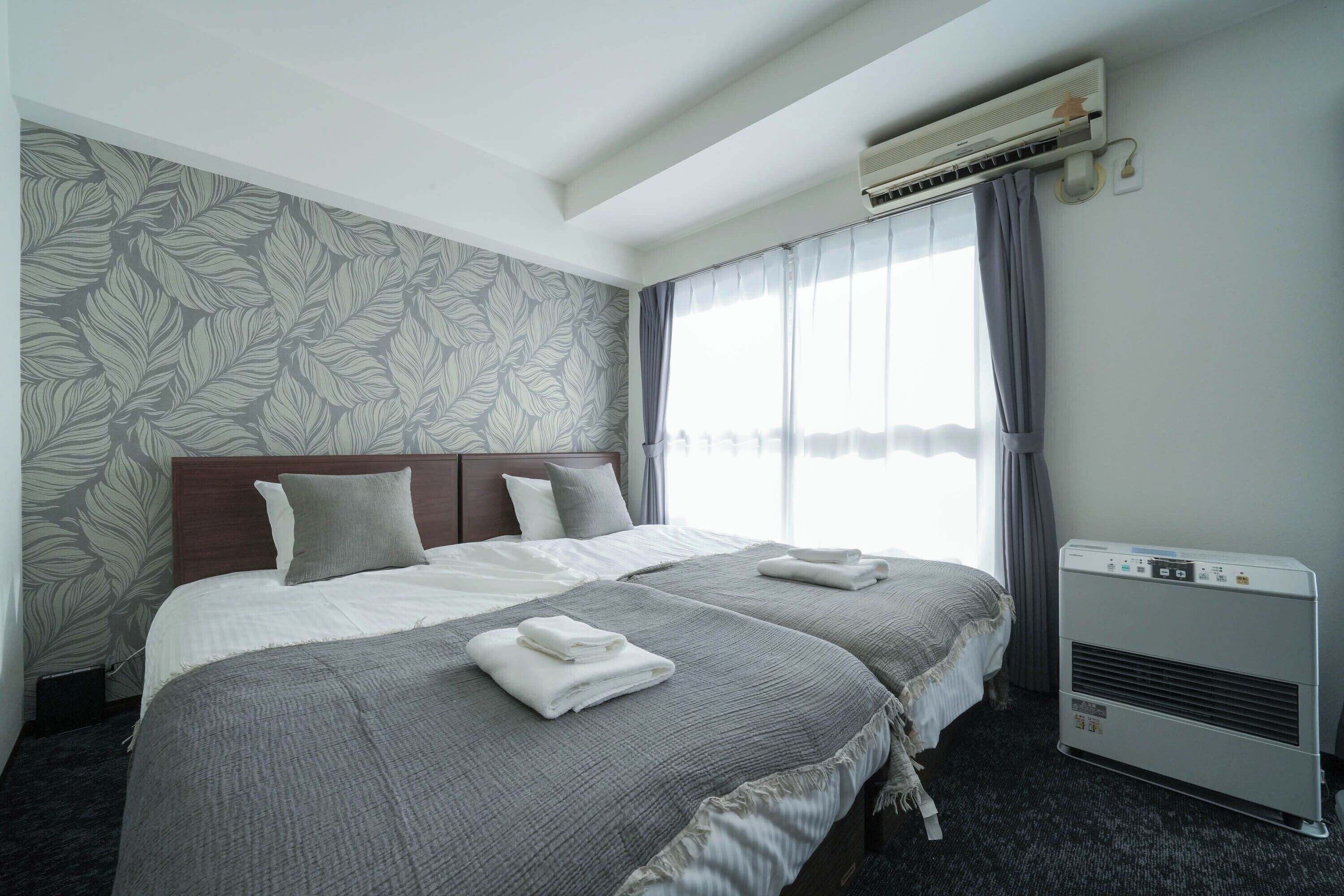 Une chambre d’hôtel avec deux lits, une grande fenêtre avec des rideaux, un appareil de climatisation et un papier peint à motifs sur le mur.