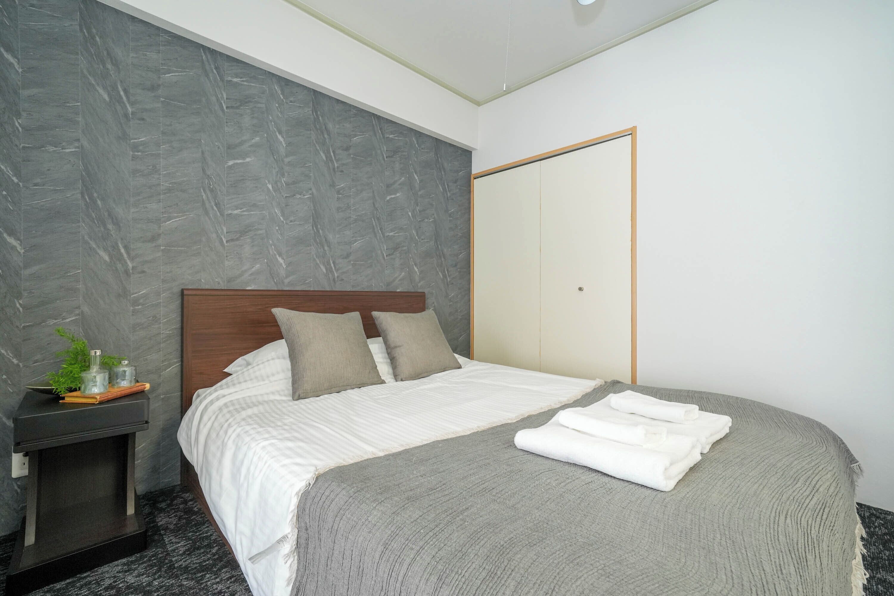 A modern hotel room with a grey textured wall, a wooden headboard, a neatly made bed with white linens, and a small bedside table with a plant and a glass.
