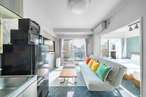 A modern hotel room with a kitchenette, a sofa with colorful cushions, a small wooden table, and a large window offering a city view. - 88 Matsui Building (Sapporo)