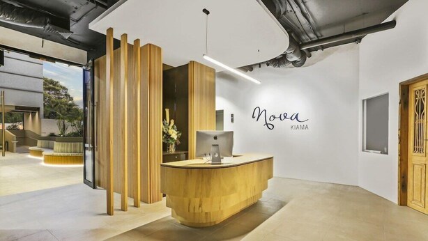 A reception desk with a wooden finish, a wall-mounted sign reading "Nova KIAMA," and a modern interior design with a visible staircase and a plant. - Nova Kiama (Wollongong)