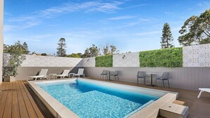 A rectangular swimming pool with clear blue water, surrounded by a wooden deck. There are lounge chairs and a small table on the deck. - Nova Kiama (Wollongong)