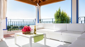 Un balcón con un sofá blanco, una mesa con una botella y flores, y vistas al paisaje.