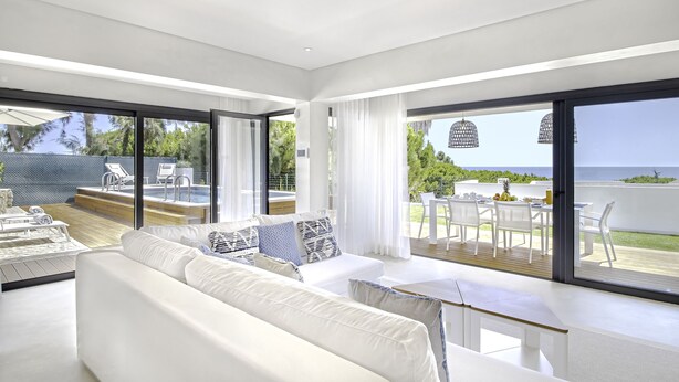 Un salón moderno con un sofá blanco, una mesa de comedor y grandes ventanales con vistas a una piscina y al océano.