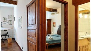 Een kamer met een houten deur, een bed met een blauw beddegoed, een houten vloer, een kleine tafel met een lamp en een wand met ingelijste foto's.