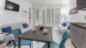 A modern, well-lit room with a grey sofa, blue cushions, a dining table with chairs, a kitchen area, and a TV mounted on the wall. - Casaviva - Val Del Ponte 5 Sanremo (Sanremo)
