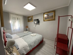 Una camera d'albergo con due letti, una TV a parete, un portariviste in metallo rosso e una finestra con tende leggere.