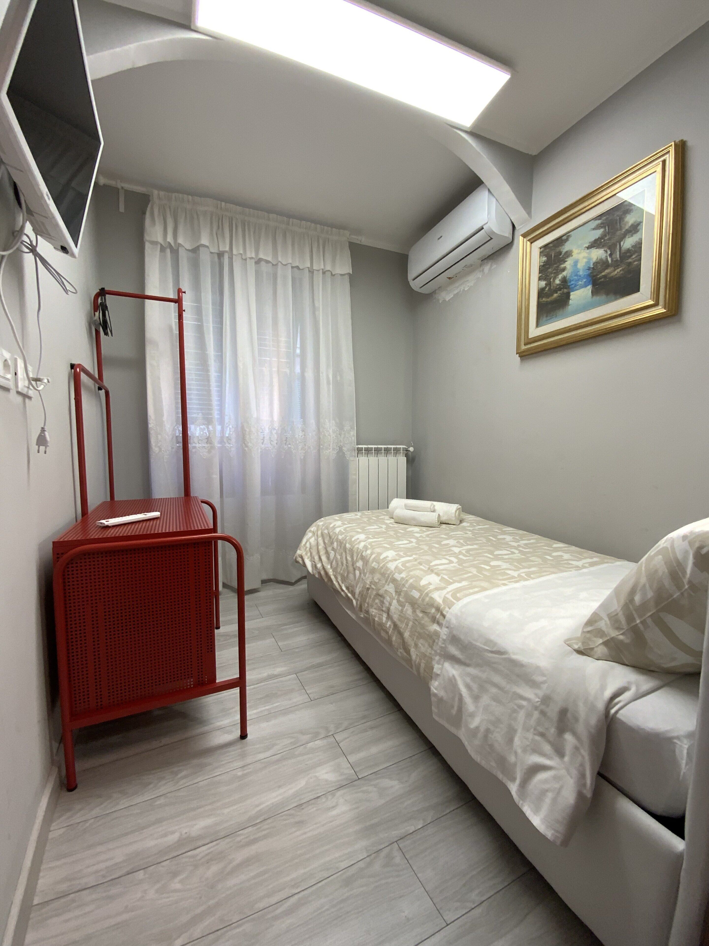 Een slaapkamer met een bed, een rode metalen stoel, een witte airconditioning en een schilderij aan de muur, en een raam met witte gordijnen.