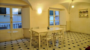 Ein Essbereich mit Tisch und Stühlen, einem kleinen runden Tisch, einem Fenster mit Jalousien und einem Regal mit dekorativen Tellern.