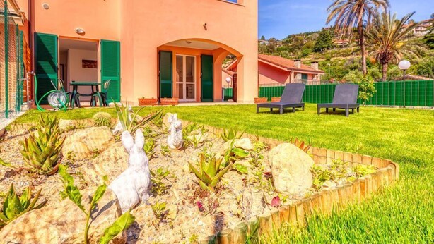 Edificio de dos pisos con un jardín al frente, que presenta una escultura de conejo blanco, plantas verdes y una valla de madera.