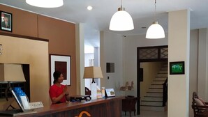 A reception desk with a receptionist, a reception area with a lamp, a framed picture, and a staircase leading to another floor. - Samawa Transit Hotel (Sumbawa Besar)