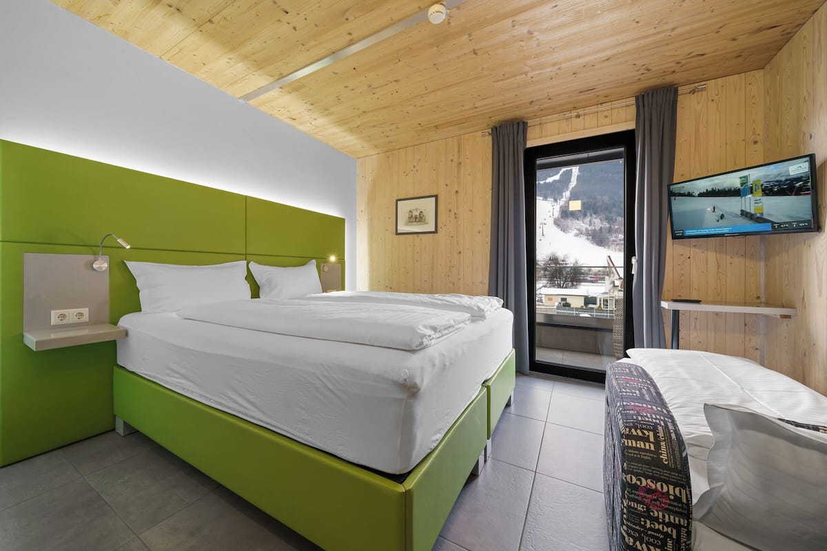 A modern hotel room with a green accent wall, a bed with white linens, a mounted TV, and a view of a snowy landscape.