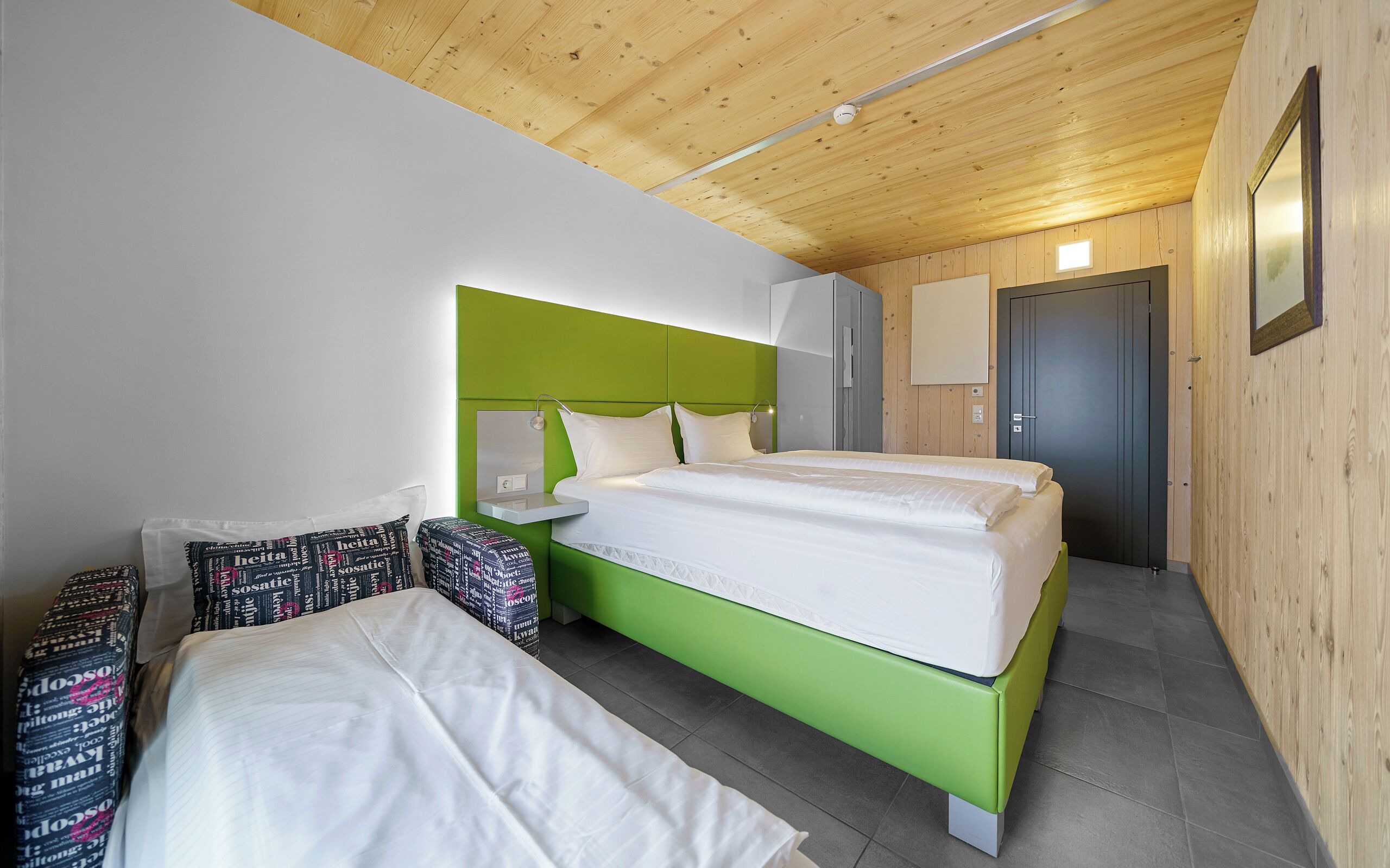 A room with a bed, a bedside table, a mirror, a door, and a wall-mounted light fixture.