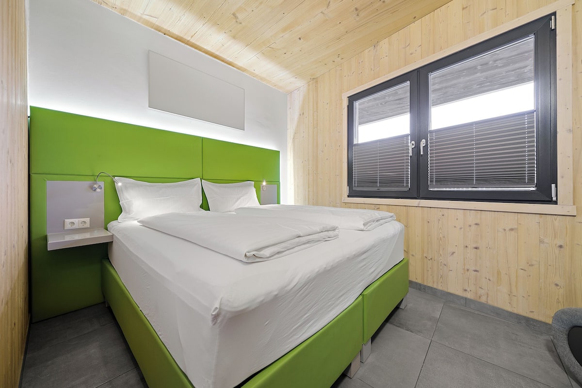 A neatly made bed in a room with a green headboard, a window with blinds, and a wooden ceiling.