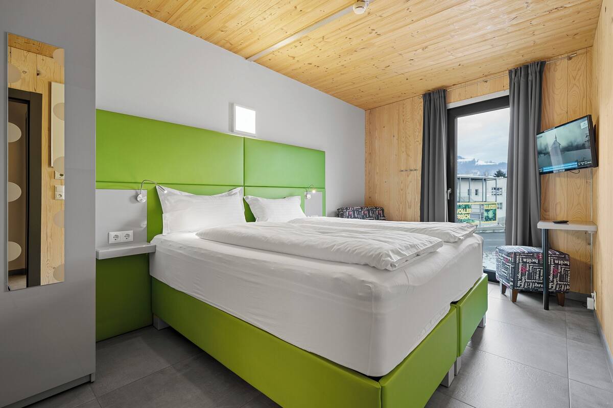 A modern hotel room with a green headboard, a large bed, a TV on the wall, and a view of the outside.